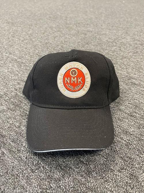 Svart caps med NMK-logo i midten, plassert på en grå teppeoverflate.