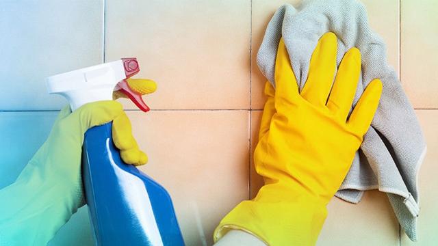 Hånd med gule gummihansker som tørker fliser med en klut, mens en sprayflaske står ved siden av.