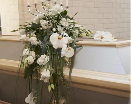 En vakker blomsterdekorasjon med hvite blomster og grønt, plassert på en bordkant i et lyst rom.