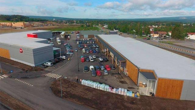 Oversiktsbilde av et kjøpesenter med parkerte biler og nærliggende bygninger under en blå himmel.