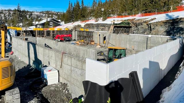 Byggeplass med betongfundament, gravemaskiner og byggeutstyr under solfylt himmel. Snø og trær i bakgrunnen.