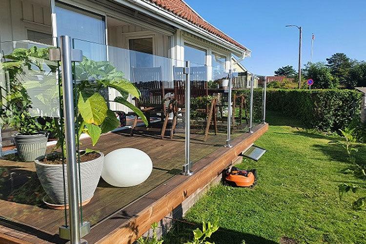 Terrasse med glassrekkverk, planter i potter og hagemøbler i solfylt hage.