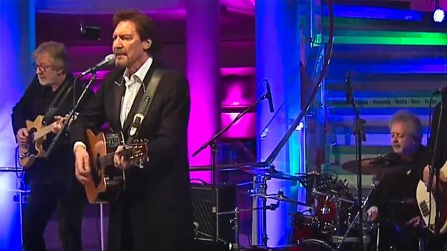 En mann synger og spiller gitar på scenen, mens to musikere spiller gitar og trommer i bakgrunnen. Fargerik belysning.