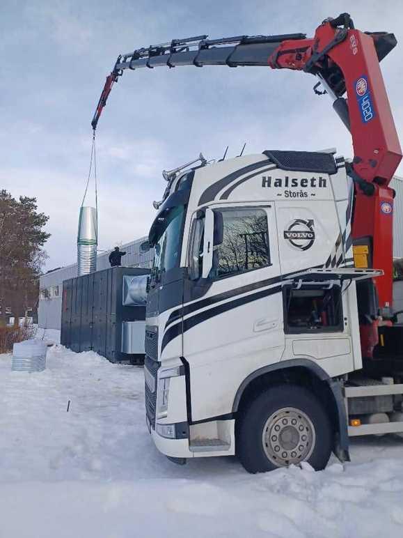 Lastebil med kran løfter en metallstruktur over en container i snødekt landskap.