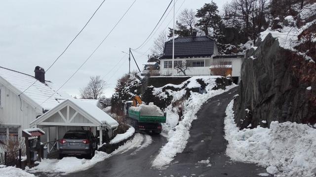 En snøfylt vei med en lastebil som rydder snø, omgitt av hus og trær i en vinterlig landskap.