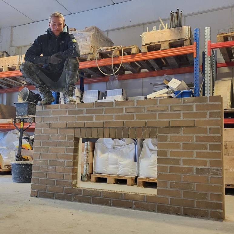 En håndverker sitter på toppen av en murvegg i et lager, omringet av byggeutstyr og materialer.