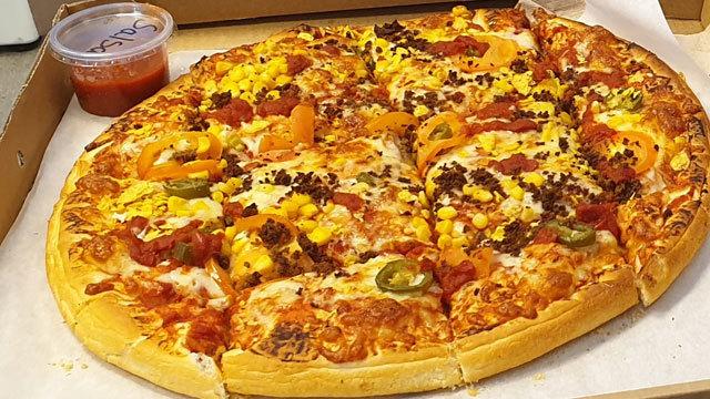 En stor pizza med ost, bacon, paprika, mais og jalapeños, servert i en pizzakartong med en liten sausbeholder.