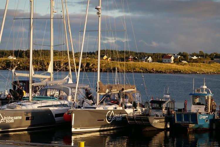 Yachter og båter ligger til kai i en havn med grønne åser og hus i bakgrunnen.