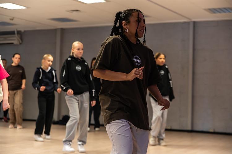 Ung kvinne danser foran en gruppe i et dansestudio, med fokus på bevegelse og rytme.