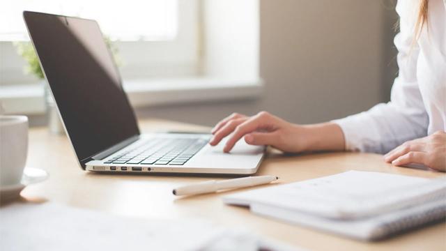 En person som skriver på en laptop på et skrivebord med en kopp og notater i bakgrunnen.