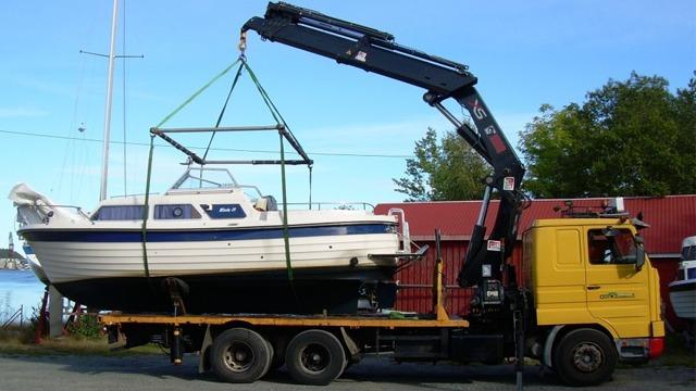 En lastebil heiser en båt med kran ved havnen på en solfylt dag.