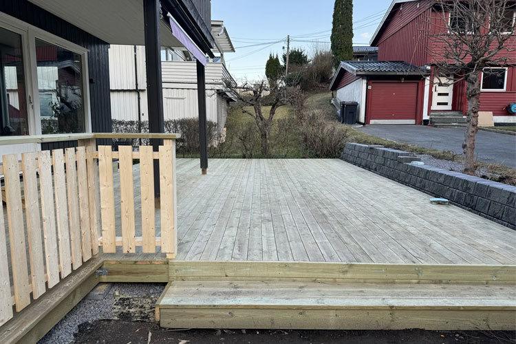 Terrasse med treplanker og rekkverk, omgitt av hage og hus i bakgrunnen.