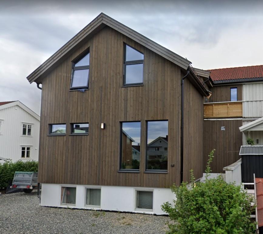 Moderne hus med trepanel og store vinduer, beliggende i et boligområde med hage og grusvei.