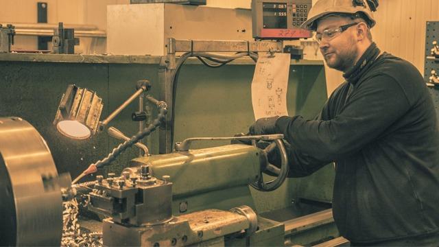 En mann som arbeider med en dreiebenk i en fabrikk, iført vernebriller og hjelm.