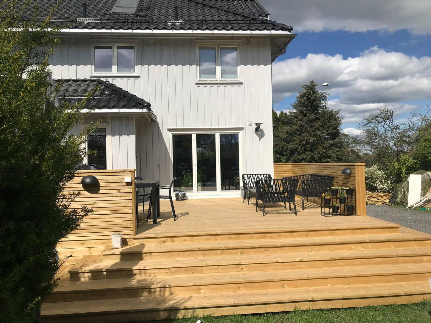 Moderne hus med stor terrasse i tre, utemøbler og grønt gress under en blå himmel med skyer.