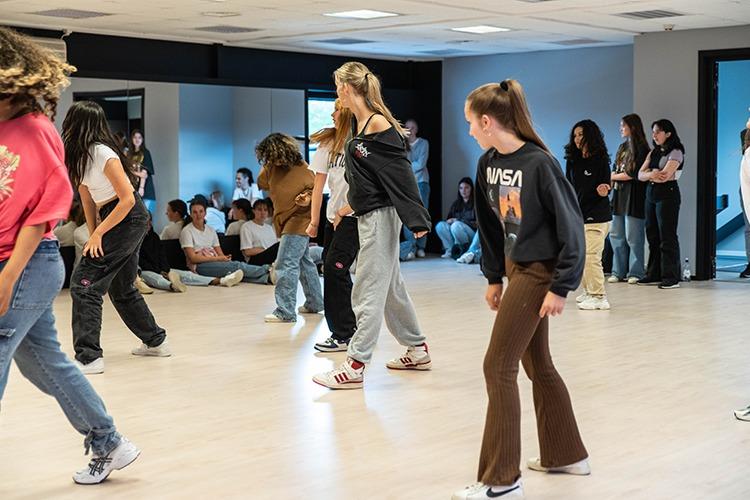 Ungdommer som danser i en lys dansestudio med speil og en gruppe som ser på.