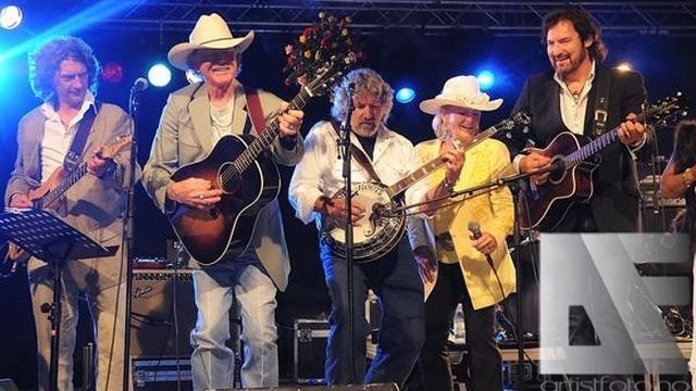 Fem musikere på scenen med gitarer og banjo, iført cowboyhatter og fargerike klær, under en konsert.