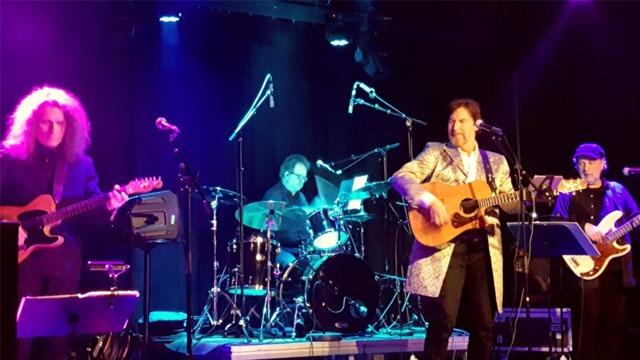 Et band som spiller live på scenen, med en sanger med gitar, trommeslager og to gitarister.