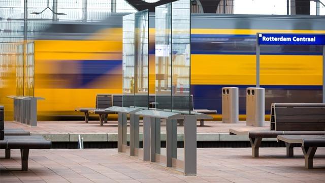 Tog som passerer på Rotterdam sentralstasjon, med benker og glasspaneler i forgrunnen.