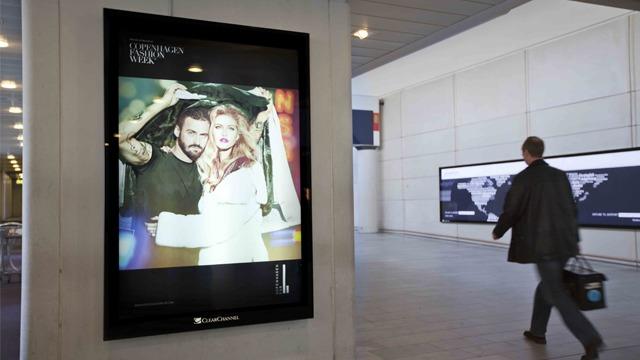 Reklameplakat med et par som poserer under en paraply, med en person som går forbi i en moderne bygning.