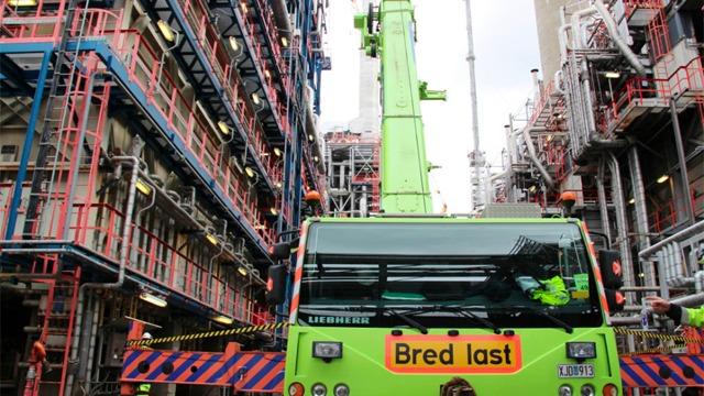 Grønn kran med teksten "Bred last" foran en industriell bygning med rør og stillas.