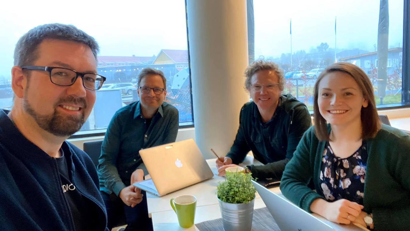 Fire personer sitter rundt et bord med bærbare datamaskiner og en plante, med vindu og utsikt i bakgrunnen.