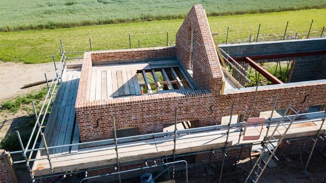 Byggeplass med murstein og takstoler under konstruksjon, omringet av stillas og grønt landskap.
