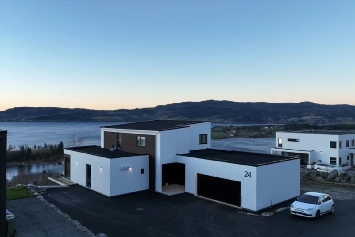 Moderne hus med hvit fasade og garasje, beliggende ved vannet med fjell i bakgrunnen.