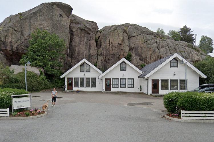 Tre hvite hus med skråtak ved en stor stein, omgitt av trær og en asfaltert vei.