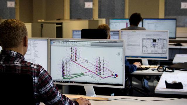 En person sitter foran en datamaskin som viser tekniske tegninger i et kontormiljø med flere skjermer.