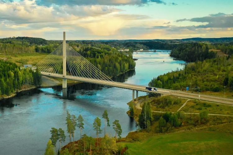 En moderne hengebro over en elv, omringet av frodig skog og landskap under en blå himmel.