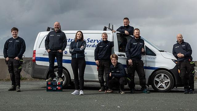 En gruppe på syv personer står foran en servicebil, iført arbeidsklær og smilende mot kameraet.