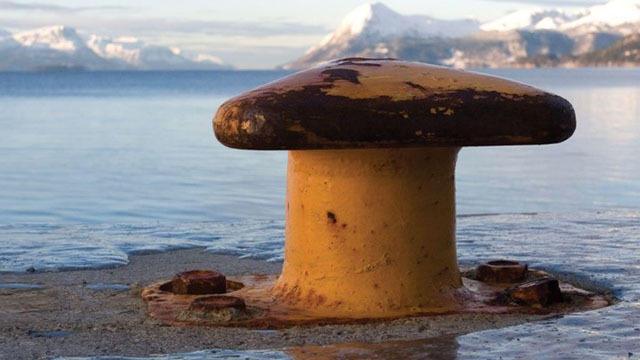 En rusten fortøyningsbøye ved vannkanten med fjell i bakgrunnen og snø på toppene.