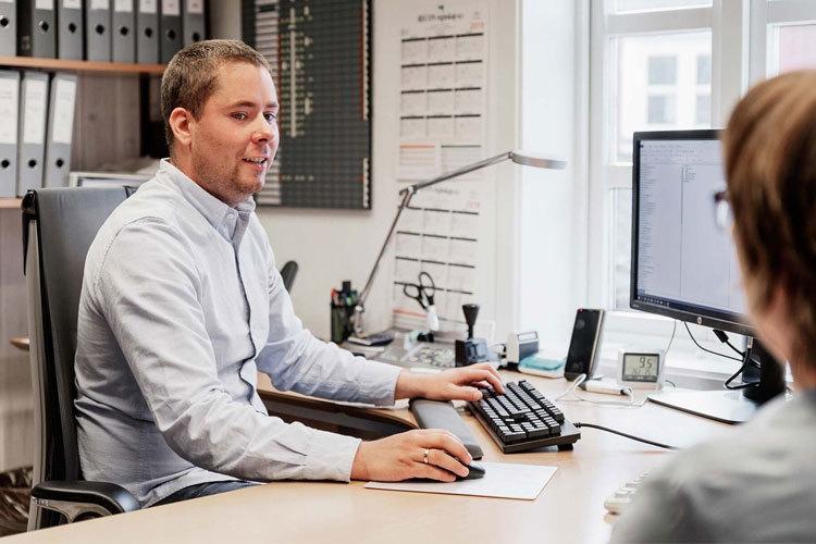 Mann i skjorte sitter ved skrivebord og jobber på datamaskin, med en kollega som ser på skjermen.