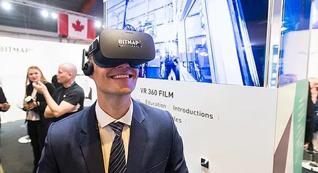 En mann i dress bruker VR-briller på en messe, med en skjerm i bakgrunnen som viser VR-innhold.