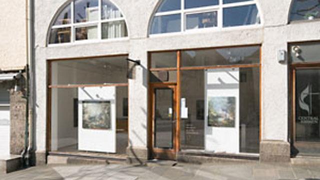 Galleri med store vinduer og utstillingsplakater på veggene, beliggende i en bygate.