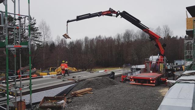 Byggeplass med kran som løfter materialer, arbeidere i sikkerhetsutstyr og byggeutstyr i bakgrunnen.