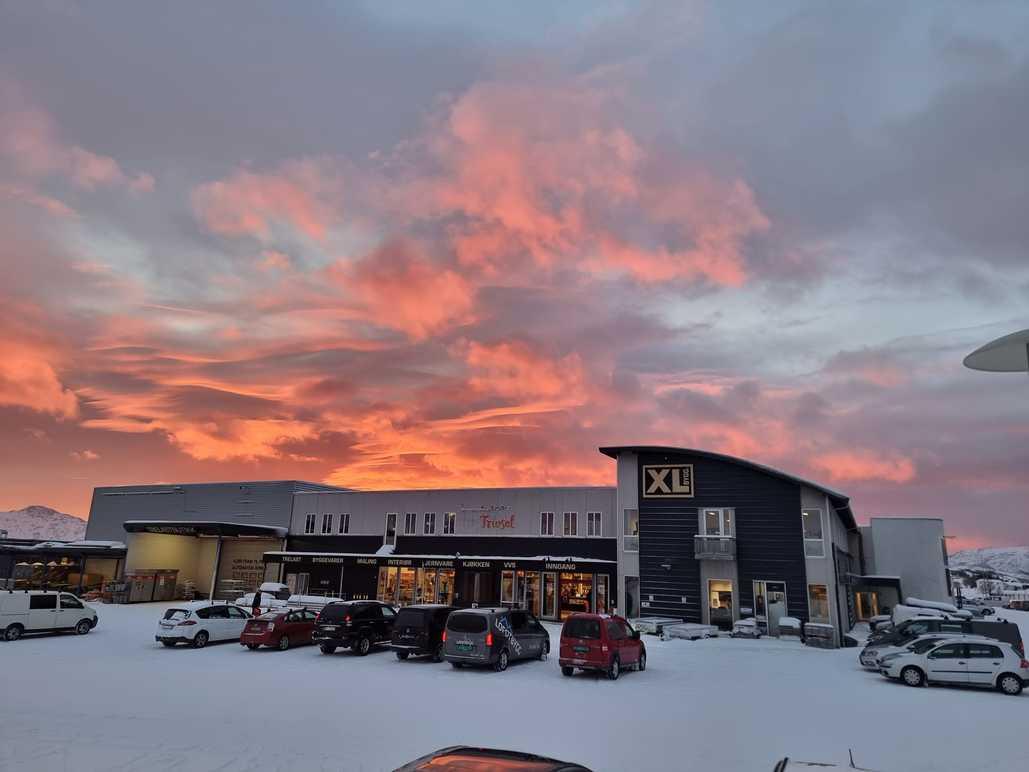 Vinterlandskap med snødekte parkeringsplasser og en butikk med fargerik solnedgang i bakgrunnen.