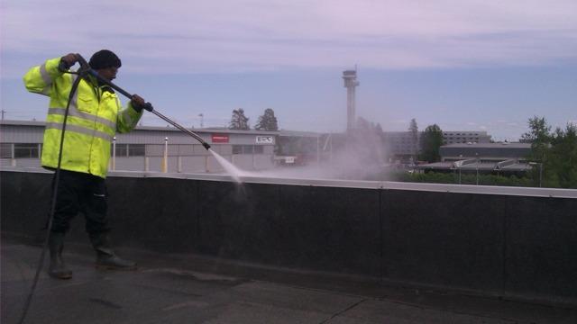 En person i refleksjakke bruker høytrykkspyler på taket av en bygning ved en flyplass.