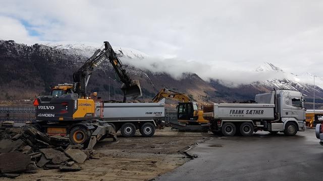 Gravemaskiner og lastebil på byggeplass med fjell i bakgrunnen og skydekke.