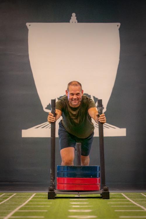 En mann trener med en sledetreningsmaskin, iført t-skjorte og shorts, på en grønn treningsmatte.