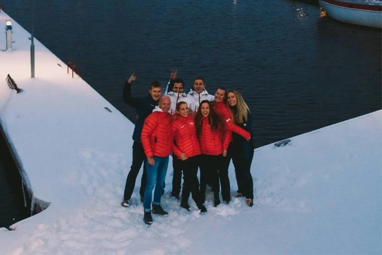 En gruppe på ni personer i røde jakker poserer smilende på snødekt bakke ved vannet.