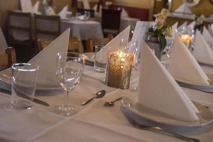 Dekket restaurantbord med hvite servietter, glass og stearinlys i en koselig atmosfære.