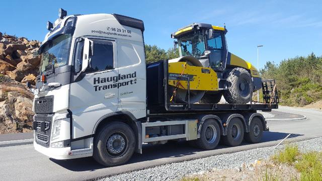 En lastebil fra Haugland Transport med en gul veivals på tilhenger, parkert langs en vei i et naturskjønt område.