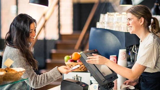 Kunde betaler med kort til en smilende ansatt i en kafé med frukt og bakverk i bakgrunnen.
