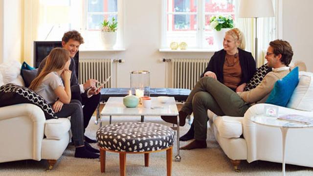 En gruppe mennesker sitter i en lys stue, og diskuterer rundt et bord med stearinlys og puter.