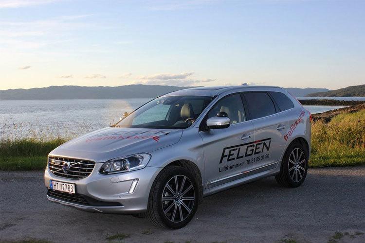En sølvfarget Volvo SUV parkert ved vannet med fjell i bakgrunnen. Bilen har påskrift fra Felgen.