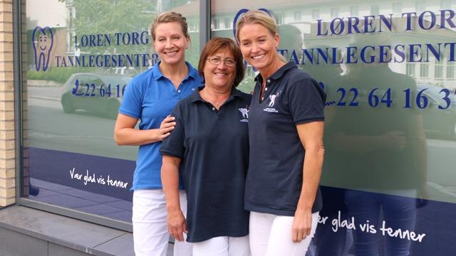 Tre tannleger står smilende foran Løren Torg Tannlegesenter, med logo og kontaktinformasjon i bakgrunnen.