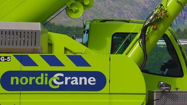 Grønn kran med "nordiCrane" logo, del av en byggeplass i naturskjønt landskap.
