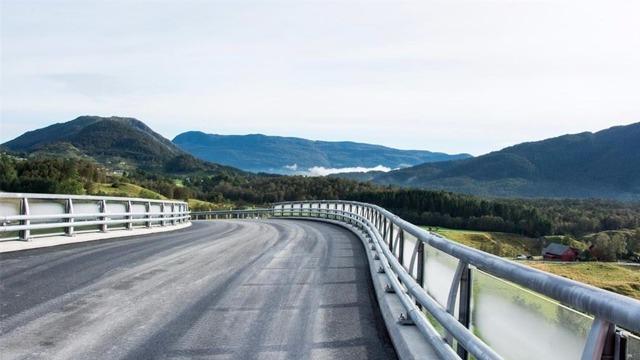 En svingete vei med rekkverk, omringet av fjell og daler under en klar himmel.